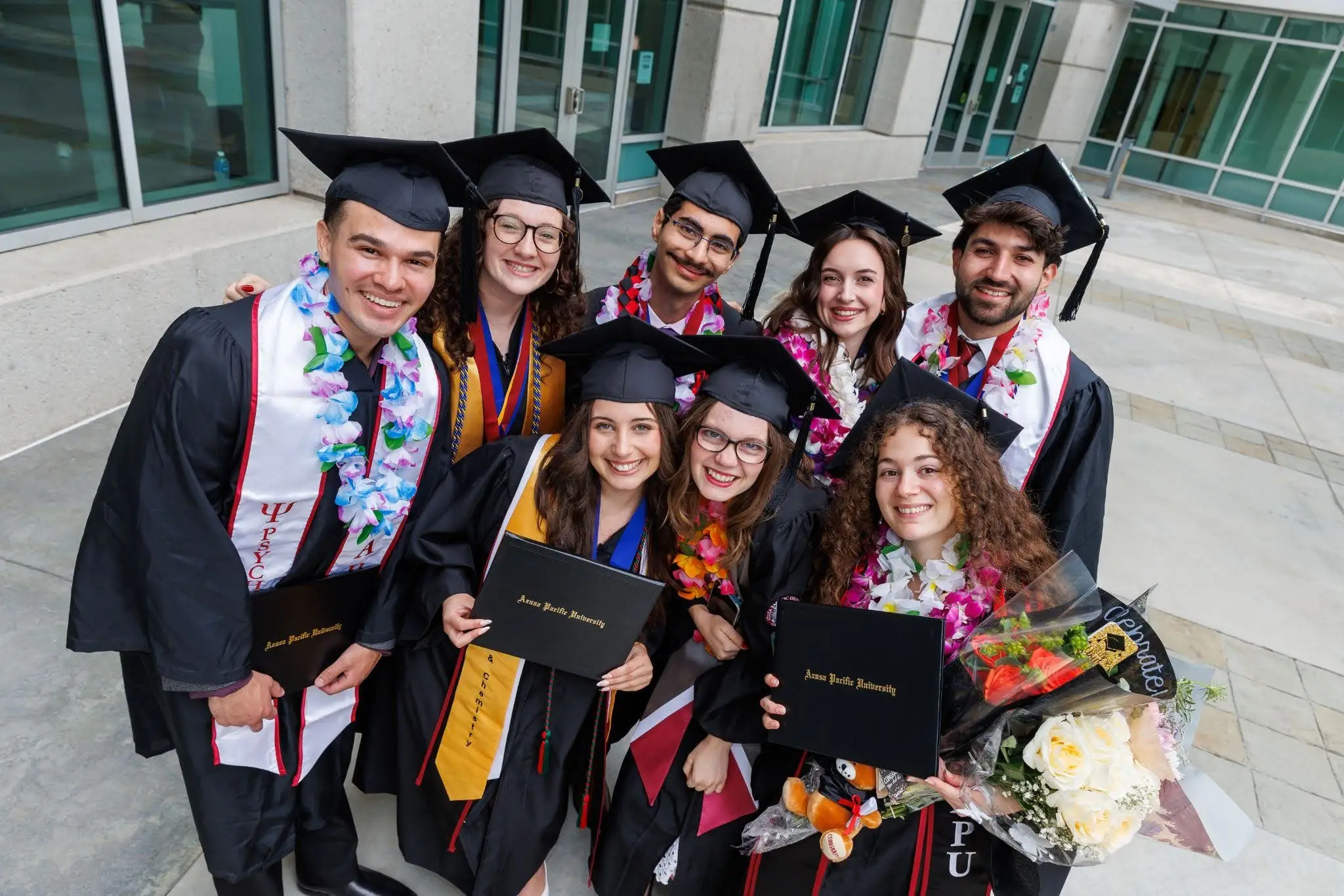 Azusa Pacific University graduates in caps and gowns pose together holding diplomas and flowers after commencement.