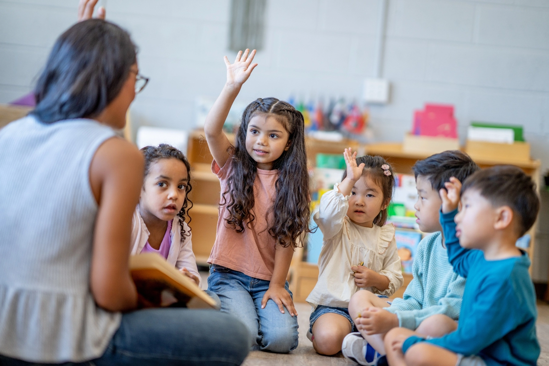 Preschool children seated in a classroom raise their hands while listening to a teacher reading a book.