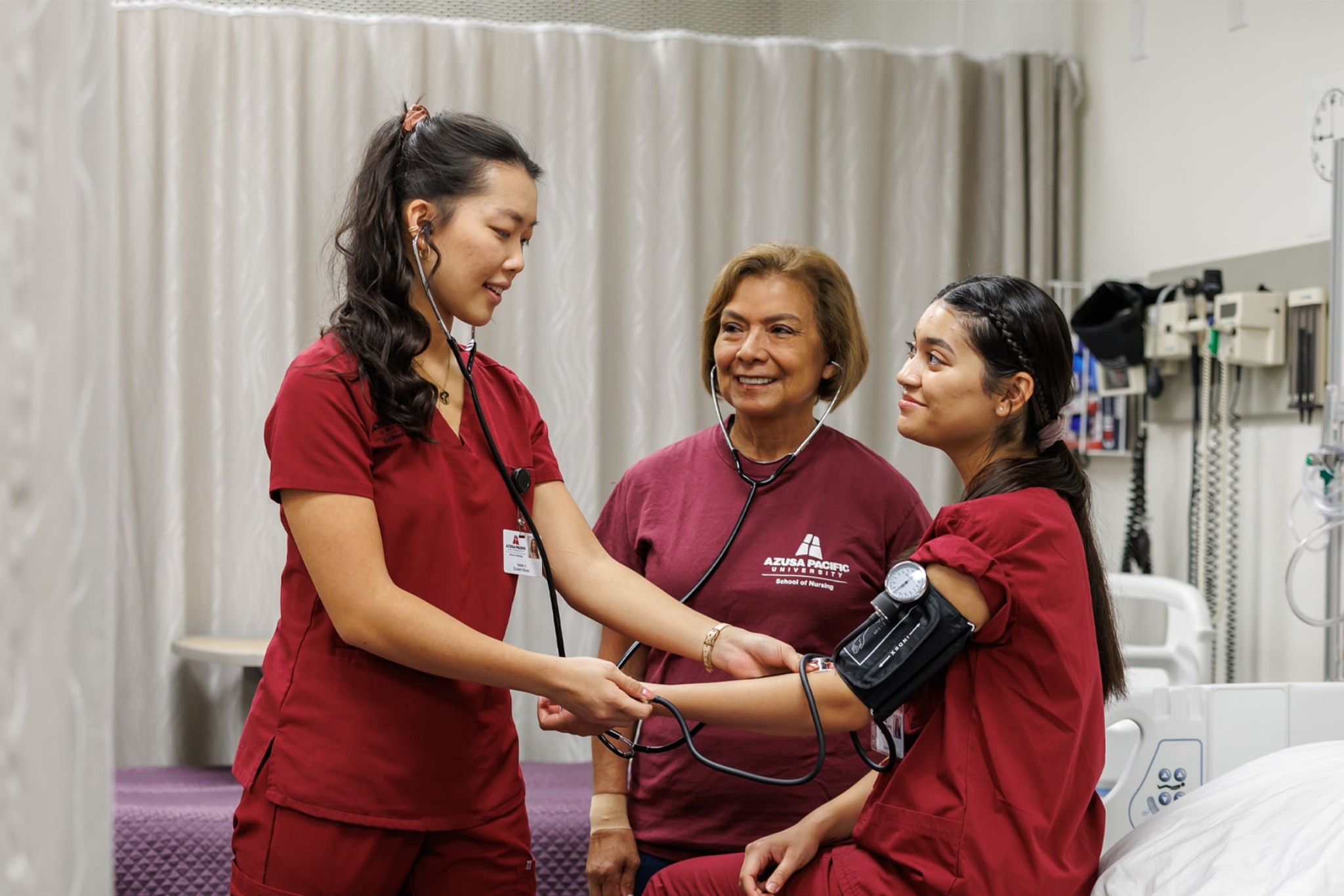 nursing students in brick red taking blood pressure of another student with their instructor guiding them