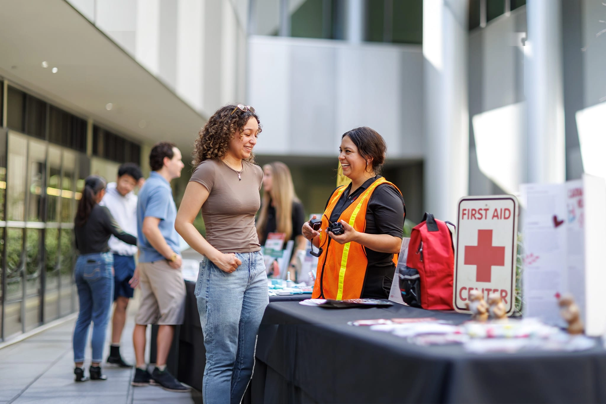 A woman in an orange safety vest smiles while speaking with a student at a First Aid Center information table during an outdoor campus event.