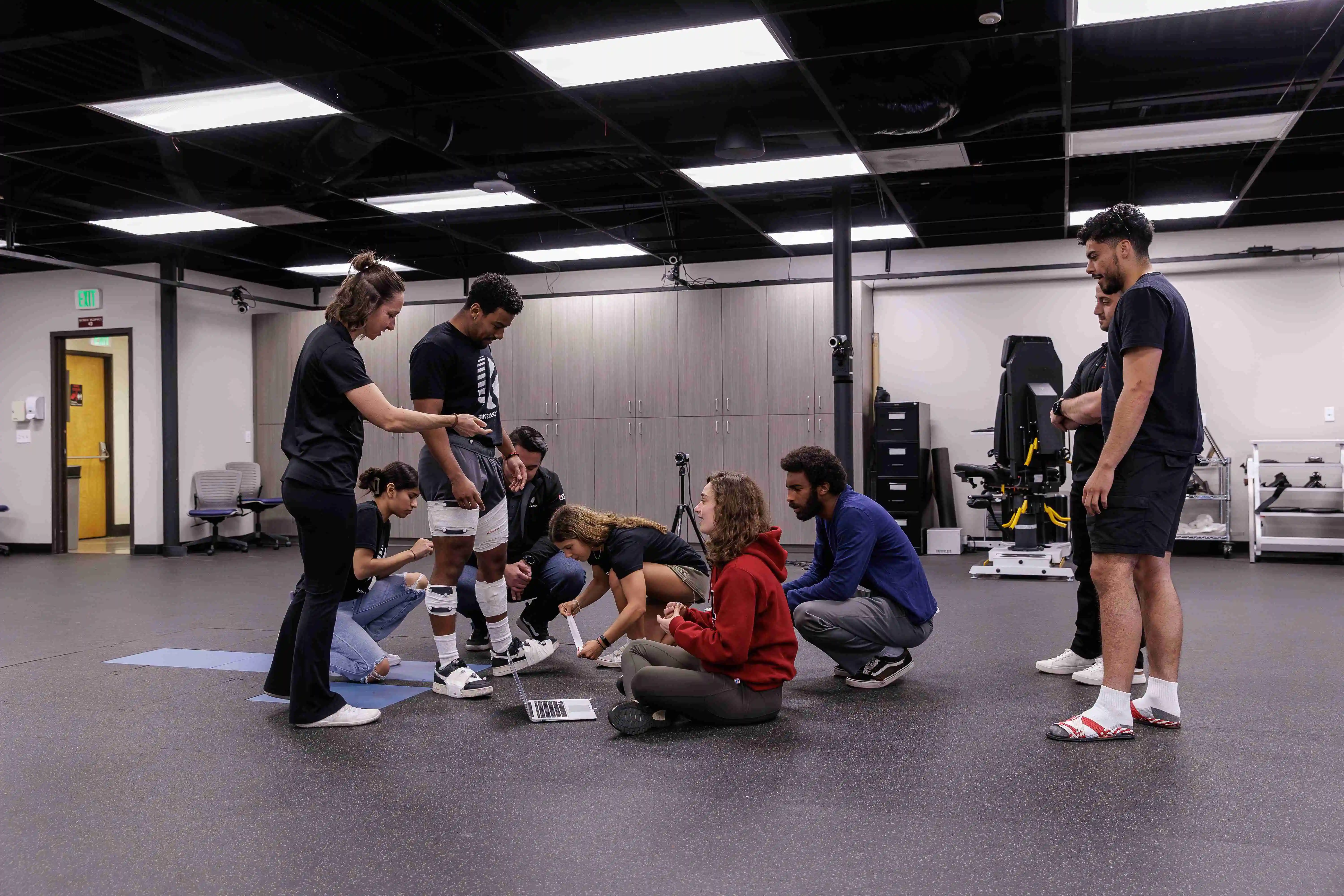Kinesiology students and an instructor in a biomechanics lab applying sensors to a subject's leg while reviewing data on a laptop.